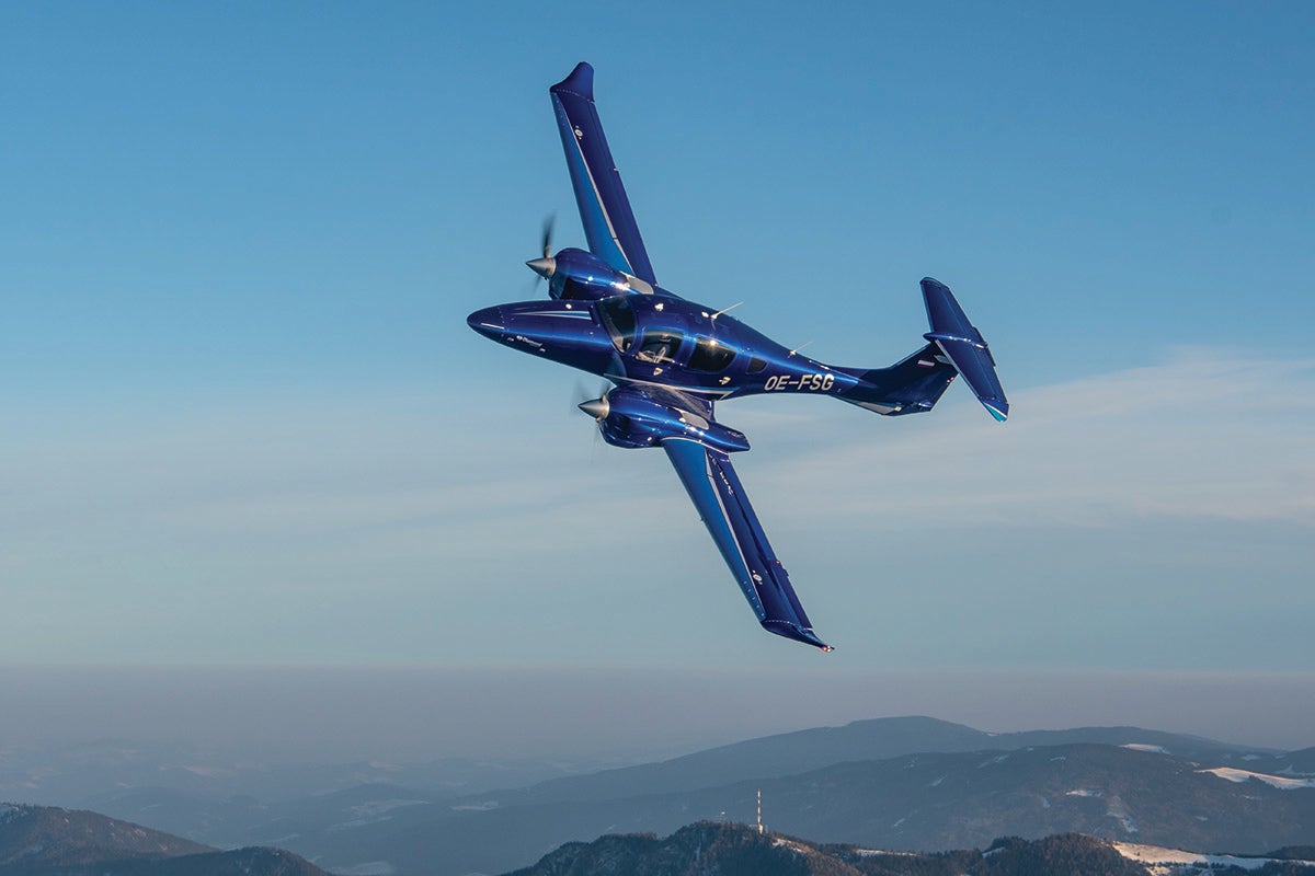 A Diamond D62 airplane soars through clear skies over low mountains in the background.