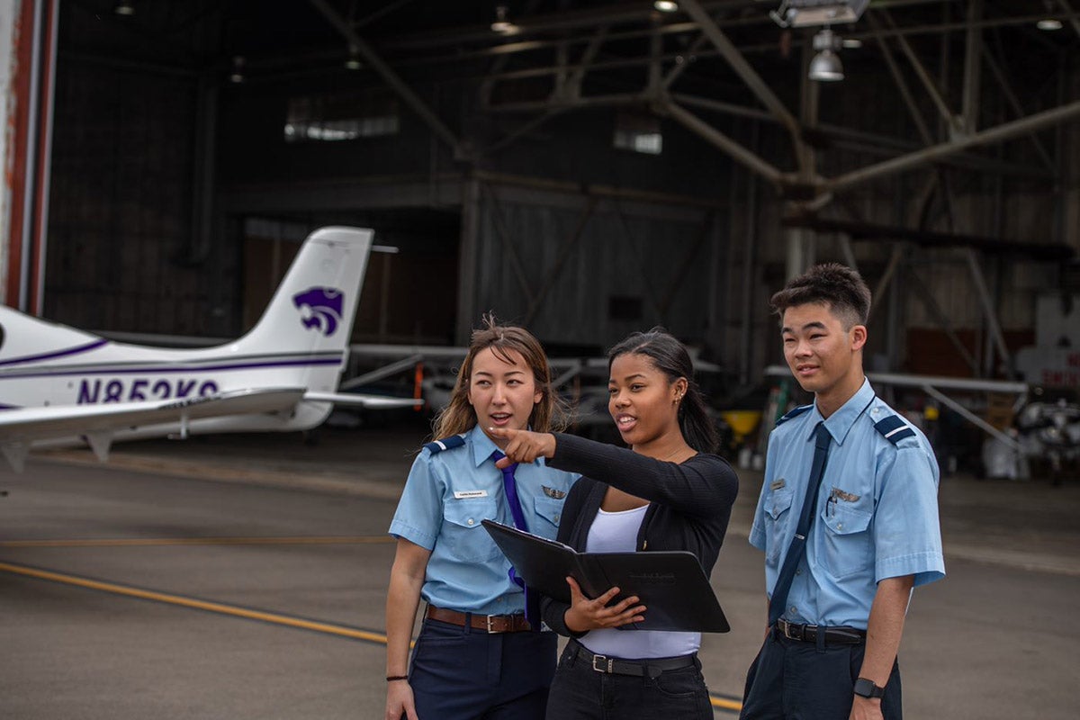 A Black female student points at the tarmac as two CFIs look on. A training aircraft and a hangar are in the background
