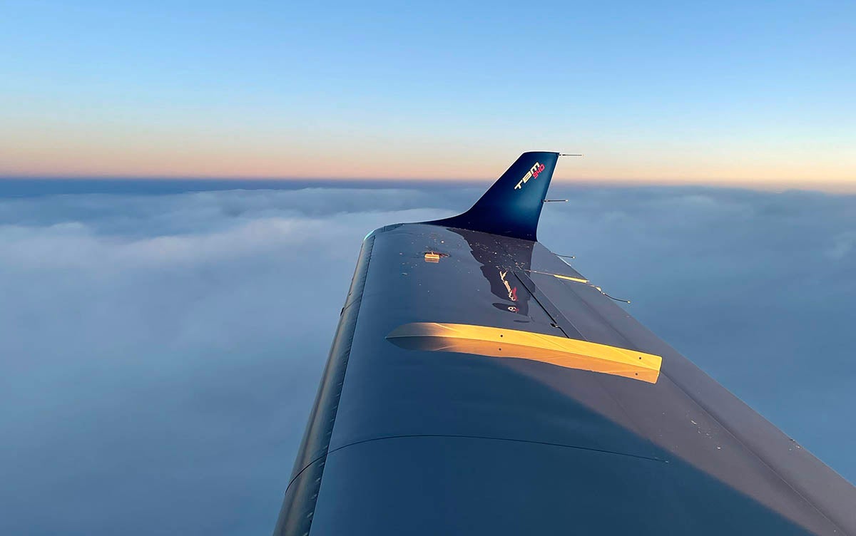 The wing and wingtip of a TBM 940 aircraft with a bed of clouds below and the horizon in the distance