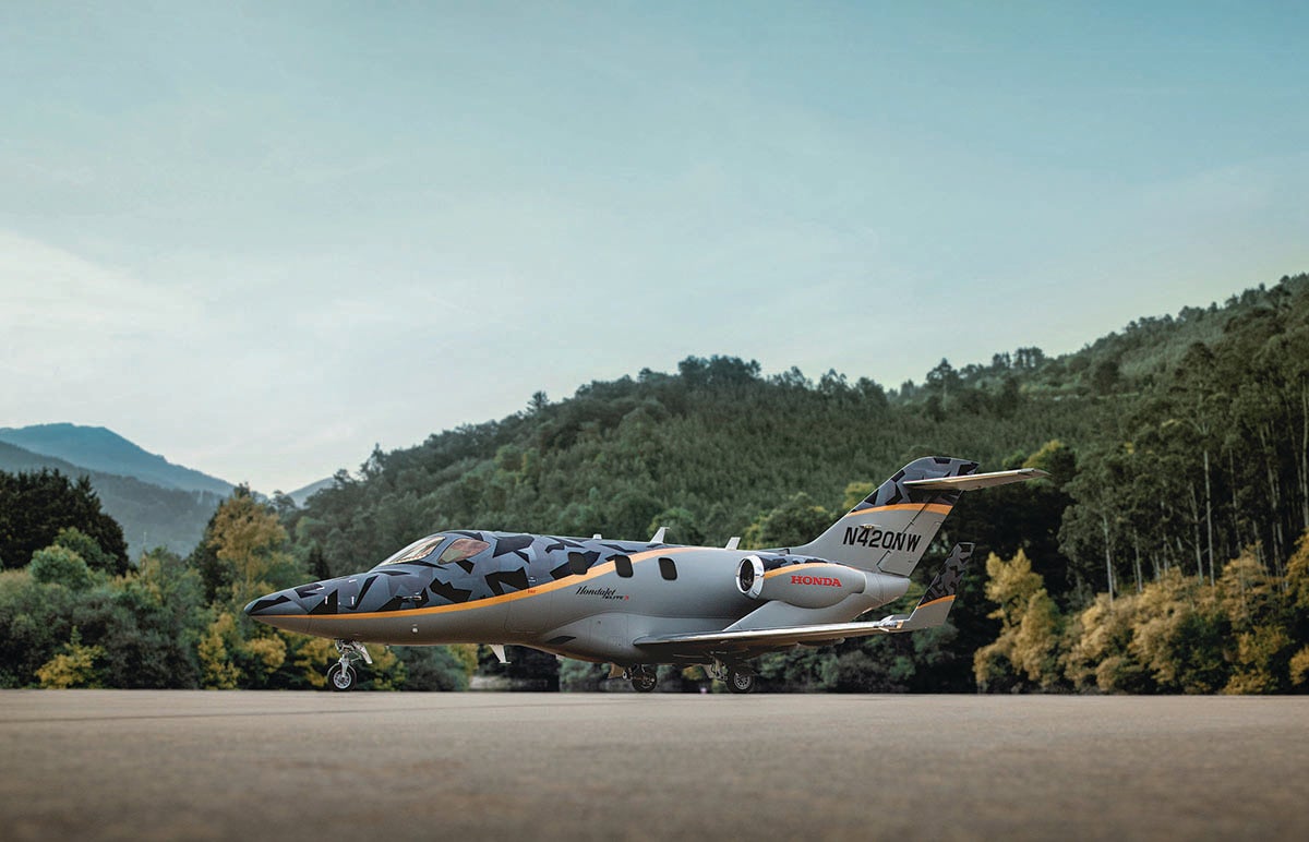 A HondaJet Elite S airplane in a gray camouflage livery sits on a tarmac with green trees and hills in the background